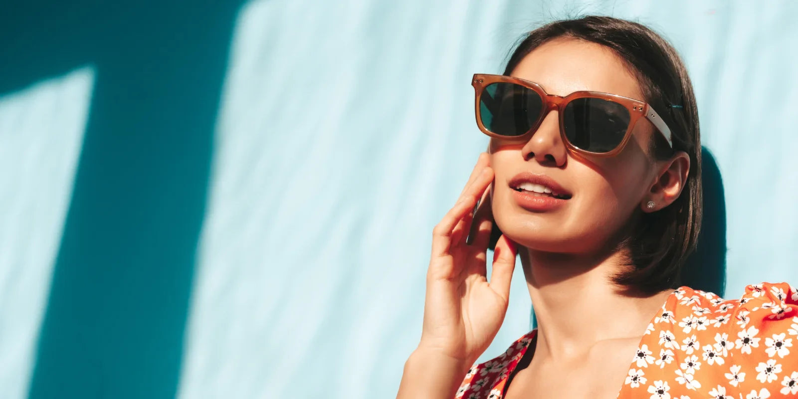 Woman in stylish sunglasses and red floral dress standing against a blue wall, showcasing trendy summer eyewear essential for Indian summers